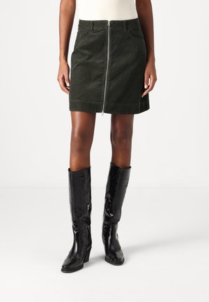 Kvinde iført en mørkegrøn cordnoy mini-nederdel med lynlås foran og sorte knæhøje støvler med krokodillemønster, stående mod en lys baggrund.