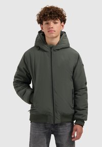 Tiener jongen met krullend haar in een groene winterjas met capuchon en grijze spijkerbroek, staand tegen een lichte achtergrond.