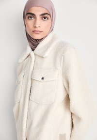 Cremefarbene, strukturierte Fleecejacke mit umschlagbarem Kragen, zwei Brusttaschen und einem Seitenetikett. Der Stoff hat ein weiches, plüschiges Aussehen.