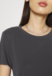 Mörkgrå t-shirt med rund halsringning och korta ärmar, bärs med flera lager av silverhalsband i olika stilar och texturer.