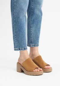 Sandali in suede marrone a scivolo con punta aperta e una larga fascia superiore. Soletta spessa color beige con superficie testurizzata. Indossati con jeans blu a vita alta.
