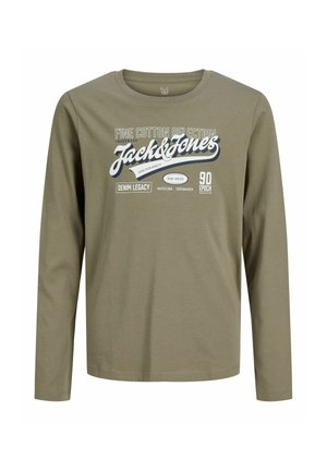 Nastolatek z kręconymi włosami, ubrany w zieloną bluzkę z długim rękawem Jack & Jones, luźne niebieskie dżinsy i brązowe buty, stoi na białym tle, patrząc na bok.
