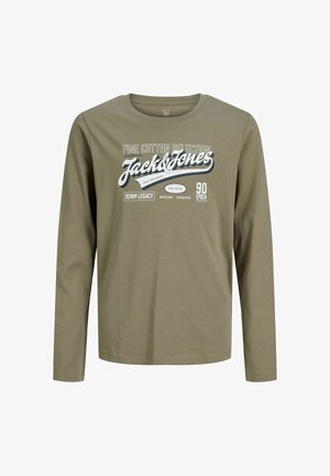 Nastolatek z kręconymi włosami, ubrany w zieloną bluzkę z długim rękawem Jack & Jones, luźne niebieskie dżinsy i brązowe buty, stoi na białym tle, patrząc na bok.