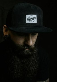 Black corduroy cap with a white rectangular patch labeled "Blackskies." The wearer has a closely trimmed beard and facial piercings.