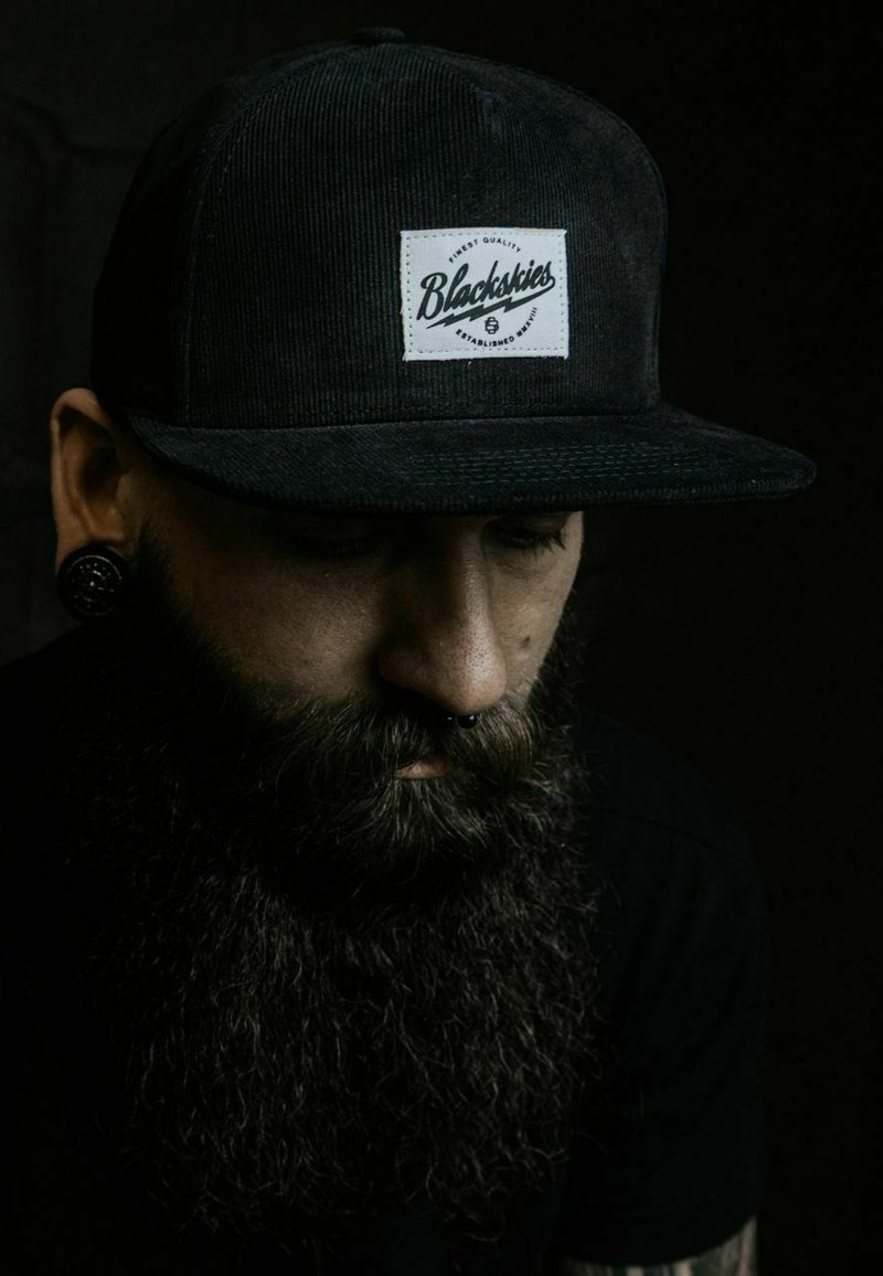 Black corduroy cap with a white rectangular patch labeled "Blackskies." The wearer has a closely trimmed beard and facial piercings.