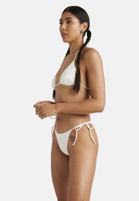 Bikini set s bielym pozadím a kvetinovými vzormi. Trojuholníkový vrch s tenkými ramienkami a nohavičky s bočnými viazaniami a nízkym strihom.