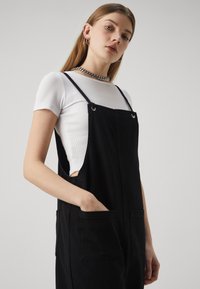 Svarte dungarees laget av slitesterkt stoff, med to frontlommer, justerbare stropper og en avslappet passform over en hvit ribbestrikket T-skjorte.