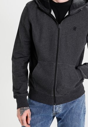 Sudadera con cremallera de color gris oscuro, con capucha, bolsillos frontales y logo negro en el pecho. Presenta una tela suave y puños y dobladillo acanalados.
