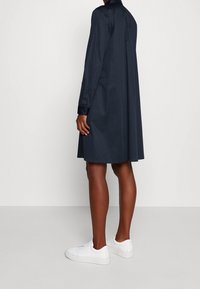 Tmavě modré šaty košilového střihu vyrobené z hladké tkaniny, s dlouhými rukávy, límcem a volným střihem, doplněné o bílé tenisky.