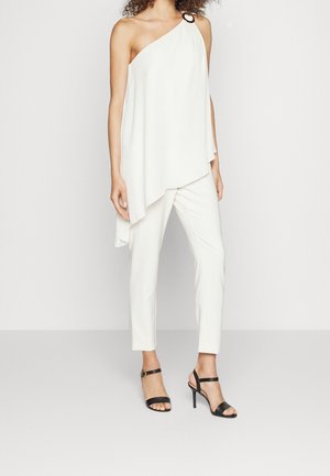 Femme portant un haut asymétrique blanc à une épaule avec un détail anneau, un pantalon blanc slim, et des sandales à talons hauts noires, debout devant un fond uni.