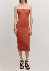 Mouwloze geribbelde jurk in roestkleur, aansluitende silhouet, knie-lengte. Gecombineerd met bruine plateau-sandalen met kruisbanden. Gouden armband.
