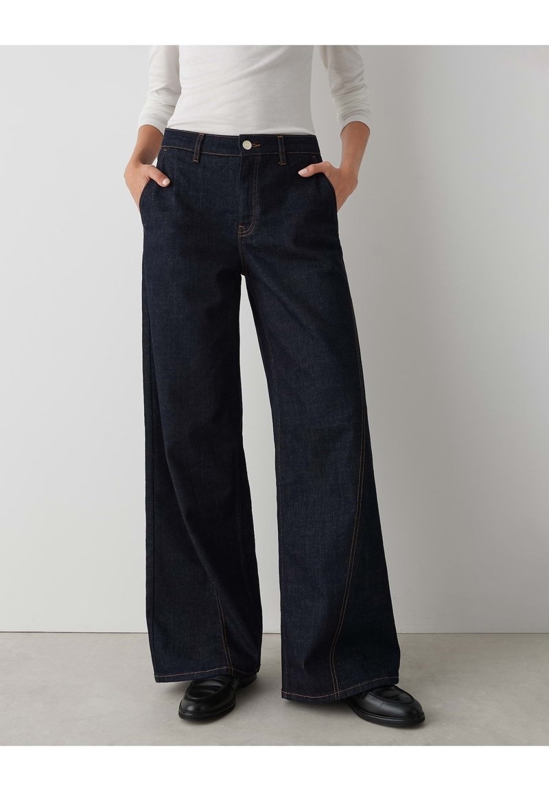 Personne portant un jean bleu foncé à jambes larges, un haut blanc à manches longues et des chaussures noires, debout, les mains dans les poches, sur un fond uni.
