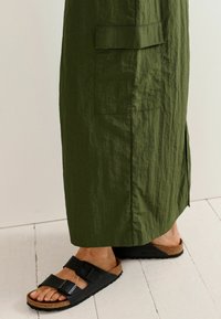 Rochie verde măsliniu, până la genunchi, din țesătură texturată cu slits pe laterale. Sandale negre cu talpă din plută și catarame cu două curele.