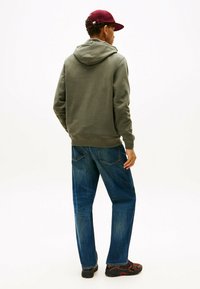 Felpa verde oliva con cappuccio, abbinata a jeans blu larghi e scarpe nere con accenti rossi. Sfondo bianco semplice.