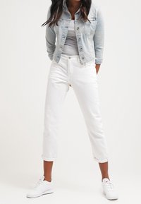 Donna con giacca di jeans chiara, top grigio, jeans bianchi arrotolati e sneakers bianche in piedi con le mani dietro la schiena.