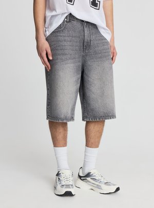 Osoba ubrana w szare dżinsowe krótkie spodenki do kolan, białe sportowe skarpetki, białe i szare sportowe buty oraz luźną białą siateczkową koszulkę.