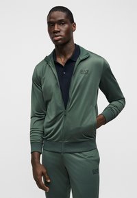 Πράσινο αθλητικό μπουφάν με φερμουάρ, με λείο υφή και ριμπ μανσέτες, συνδυασμένο με μαύρο πόλο μπλουζάκι που φέρει ένα μικρό λογότυπο στο στήθος.