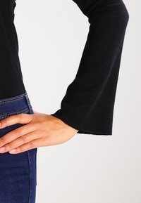 Czarna bluzka z długim rękawem o prążkowanej strukturze i rozszerzonym mankiecie, w zestawieniu z ciemnoniebieskimi dżinsami. Ręka spoczywa na pasku.