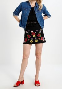 Giacca di jeans sopra un vestito nero con ricami floreali, abbinato a sandali a tacco quadrato rossi. La gonna presenta vivaci motivi floreali in più colori.