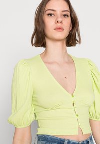 Blusa curta verde-clara com mangas bufantes, apresentando um decote em V profundo e seis botões decorativos na parte da frente. Tecido texturizado.