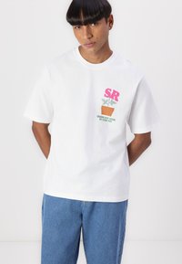 White cotton t-shirt featuring a pink "SR" logo and a green plant graphic above a terracotta pot. Slightly loose fit with short sleeves.