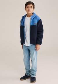 Kék és sötétkék polárkabát kapucnival, elöl cipzárral és texturált anyaggal. Fehér póló és kék farmer fölött viselve.