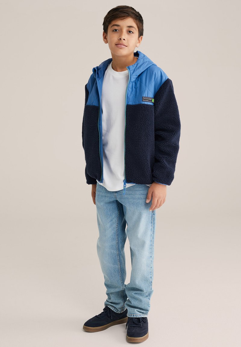 Kék és sötétkék polárkabát kapucnival, elöl cipzárral és texturált anyaggal. Fehér póló és kék farmer fölött viselve.