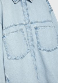 Light blue denim jacket with a pointed collar, two chest pockets, and visible stitching. The fabric has a smooth texture and slightly faded finish.