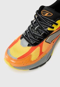Sneaker, millekollase võrkkattega, oranžide ja punaste aktsentidega, läikivate mustade detailidega, halli kummist tallaga ja mustade paeltega.