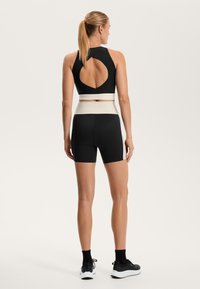 Črn pokrščen športni modrček z okroglo izrezano hrbtno stranjo, v kombinaciji s črnimi kratkimi hlačami z belim pasom. Gladka tekstura, športen dizajn.