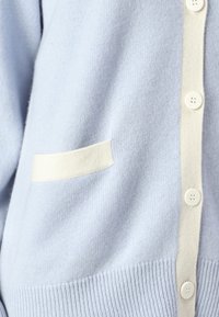 Απαλή μπλε ζακέτα από μαλακό ύφασμα, με κρεμ λεπτομέρειες, πέντε στρογγυλά κουμπιά και μια μονή θηλυκή τσέπη.