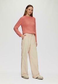 Koraalrood ribgebreid trui met lange mouwen, gecombineerd met hooggesneden, lichtbeige wijde broek en crèmekleurige chunky schoenen.