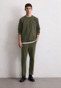 Sweatshirt vert olive avec un col rond, associé à un pantalon assorti en olive. Le modèle porte des bottes noires à lacets sur un fond neutre.
