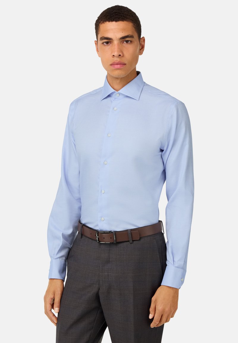 Camicetta azzurro chiaro a maniche lunghe con colletto a punte, caratterizzata da una texture sottile e una vestibilità slanciata, indossata con pantaloni grigi scuri e una cintura marrone.