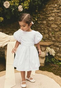 Robe en coton blanche avec des manches bouffantes, taille froncée et jupe mi-longue. Assortie à des chaussures blanches et un ruban pour les cheveux. Fond de mur en pierre.