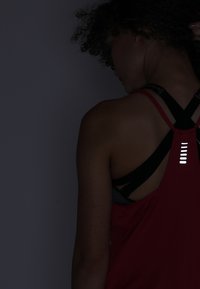 Rotes sportliches Tanktop mit überkreuzten Trägern, das schwarze Unterwäsche zeigt. Auf der Rückseite befinden sich reflektierende weiße Akzente.