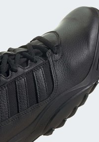 Chaussure de sport noire avec une tige en cuir lisse, des accents en maille texturée, des œillets renforcés et une semelle en caoutchouc avec des crampons de traction.