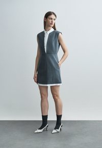 Nő ujjatlan szürke ruhában, fehér galléros ing alá húzva, fekete zoknival és fehér magassarkú cipőben, világos egyszínű háttér előtt áll.