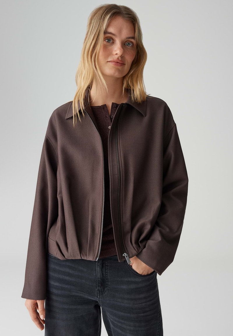 Braune Jacke mit Reißverschluss, einem Kragen und gerafftem Saum. Hergestellt aus glattem Stoff, mit langen Ärmeln und lockerem Sitz. Wird über einem dunklen Oberteil getragen.