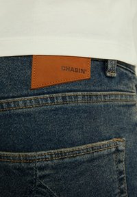 Jeans en denim bleu présentant un patch en cuir tan avec "CHASIN'" en relief, des coutures visibles et un design standard de poche arrière.