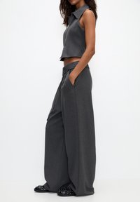 Grijze cropped mouwloze top met een kraag, gecombineerd met brede grijze broeken met zijzakken, afgewerkt met versierde zwarte schoenen.