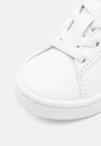 Vit sneaker tillverkad av syntetiskt material med en slät yta, med en rund tå, vita snörningar och synlig sömnad längs sömmarna.