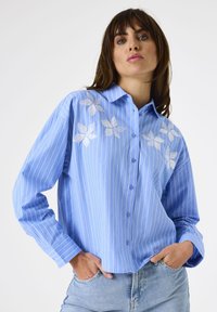 Woman wearing a light blue, long-sleeve, striped shirt with white floral embroidery paired with light blue jeans, hands in pockets.