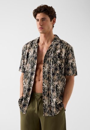 Młody mężczyzna ubrany w otwartą koszulę z krótkim rękawem w wzór palm oraz oliwkowozielone spodnie ze sznurkiem, ręce w kieszeniach, neutralne tło.