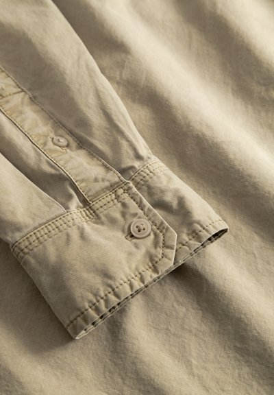 Beige button-up shirt cuff showing a stitched hem and a single button. Fabric appears soft, with a slightly wrinkled texture.