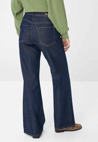 Dunkelblaue, hoch taillierte, ausgestellte Jeans mit zwei hinteren Taschen und gesteppten Akzenten. Kombiniert mit gemusterten Stiefeln und einem grünen Oberteil.