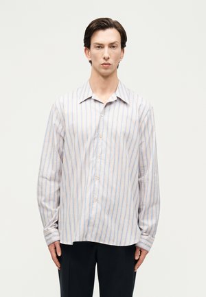 Mężczyzna ubrany w luźną, długą, zapinaną na guziki koszulę w cienkie pionowe niebieskie i pomarańczowe paski oraz czarne spodnie, stojący na gładkim tle.