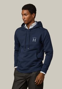 Sudadera azul marino con cordones y bolsillo frontal. Presenta un logo gris "HACKETT H" en el pecho. Superpuesta sobre una camiseta a rayas.
