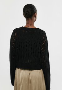 Frau mit dunklem geflochtenem Dutt, die einen schwarzen Strickpullover und einen beigen Plisseerock trägt, von hinten vor einem weißen Hintergrund betrachtet.