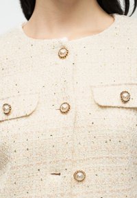 Közelkép egy krémszínű texturált dzsekit arany és gyöngy díszítőgombokkal, valamint két fedeles zsebbel a mellrészen.
